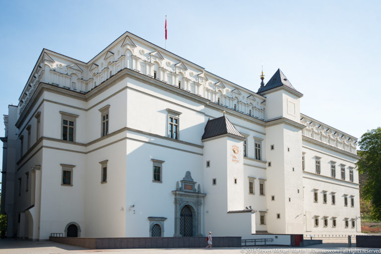 White Renaissance-style palace with regular windows, corner towers, dark roofs, stone portal, and Lithuanian flag.