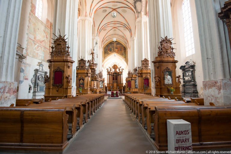 Central aisle with wooden pews, tall white piers, painted Gothic vaults, and carved altars at the sanctuary.