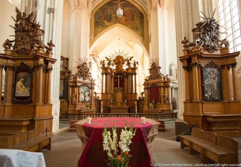 Red-covered altar table with white flowers before multiple carved wooden Baroque altars in the Bernardine Church sanctuary.