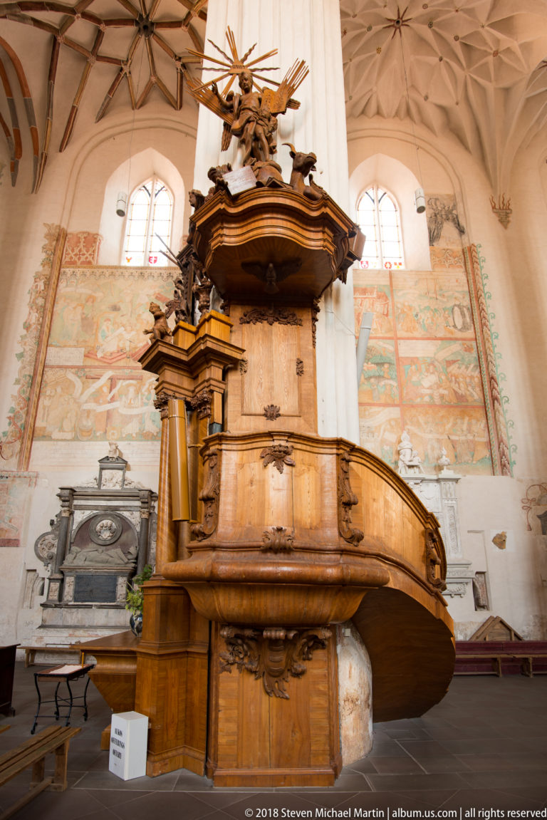 Large carved wooden pulpit with curved enclosed stair and sculptural canopy against pale wall paintings.