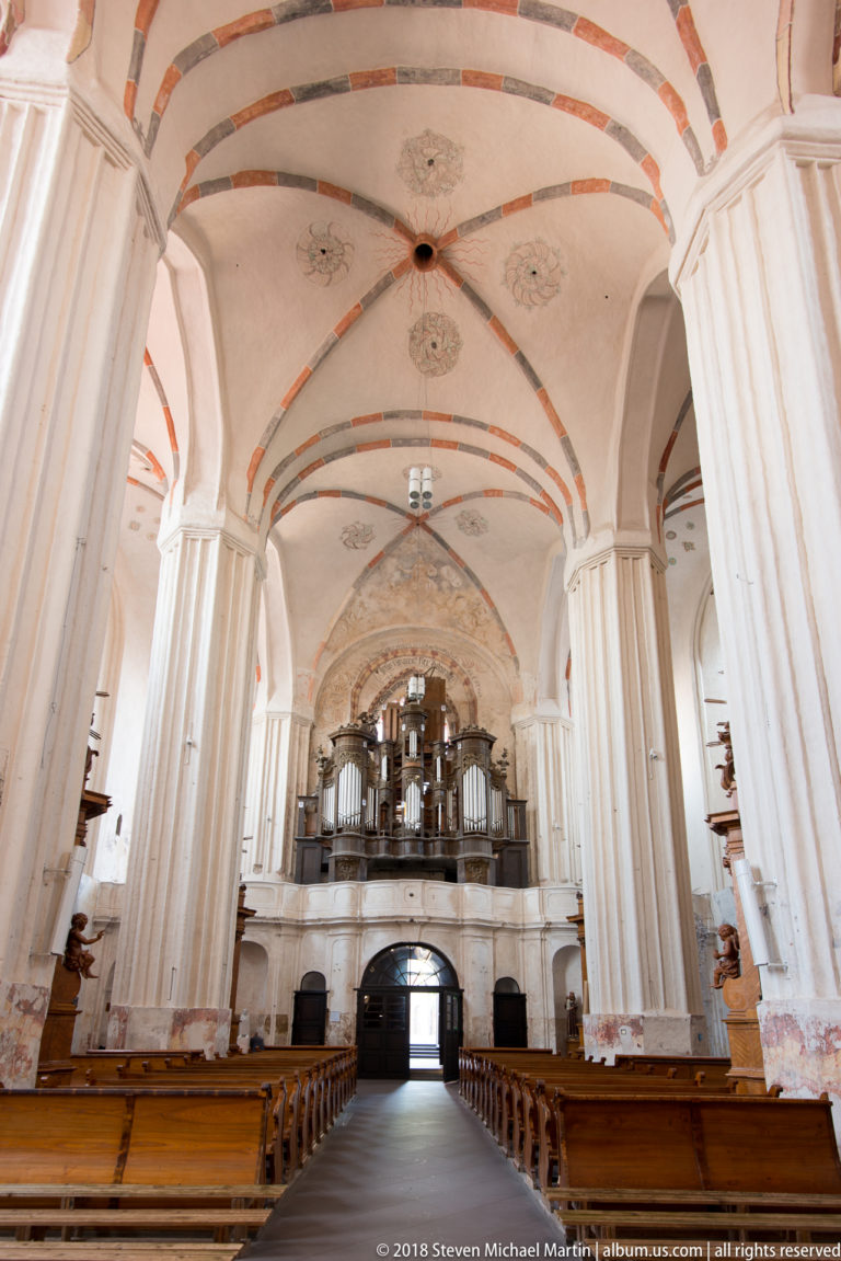 Long church nave with wooden pews, tall white piers, painted ribbed vaults, and gallery at the far end.