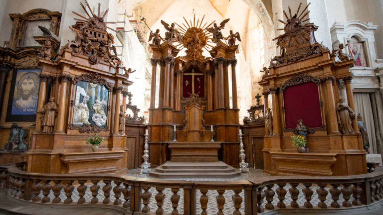 Carved wooden Baroque altar group behind a balustrade, with sacred images, red panels, and radiant sculptural crest.