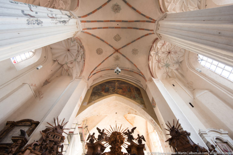 Tall white piers and ribbed Gothic vaults above carved wooden altars and faded wall paintings in the Bernardine Church.