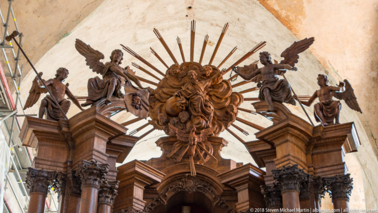 Carved wooden altar crest with central radiant figure, winged angels, sunburst rays, columns, and Baroque ornament.