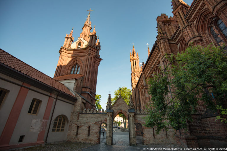 Courtyard approach with red-brick church towers, pointed gate, masonry wall, trees, and adjoining plastered building.