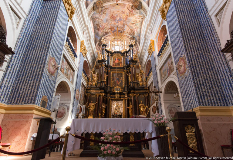 Central nave view toward an ornate dark-and-gold high altar, with blue patterned pilasters, painted vaults, chandeliers, and flowers.