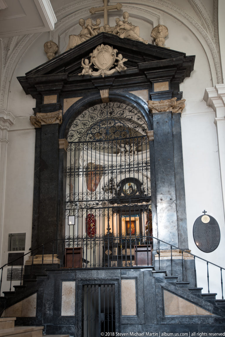 Black stone chapel portal with ornate wrought-iron grille, pale sculpted cresting, and a lit altar visible beyond.