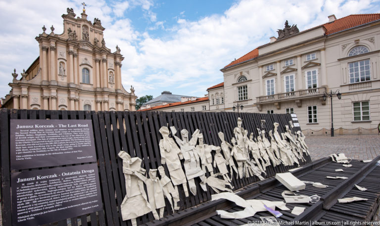 Janusz Korczak memorial display with bilingual text and a procession of white cutout figures in a Warsaw square.