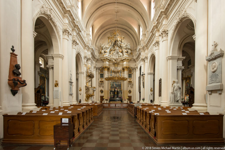 Long white church nave with wooden pews, repeated arches, wall monuments, and an ornate sanctuary in the distance.