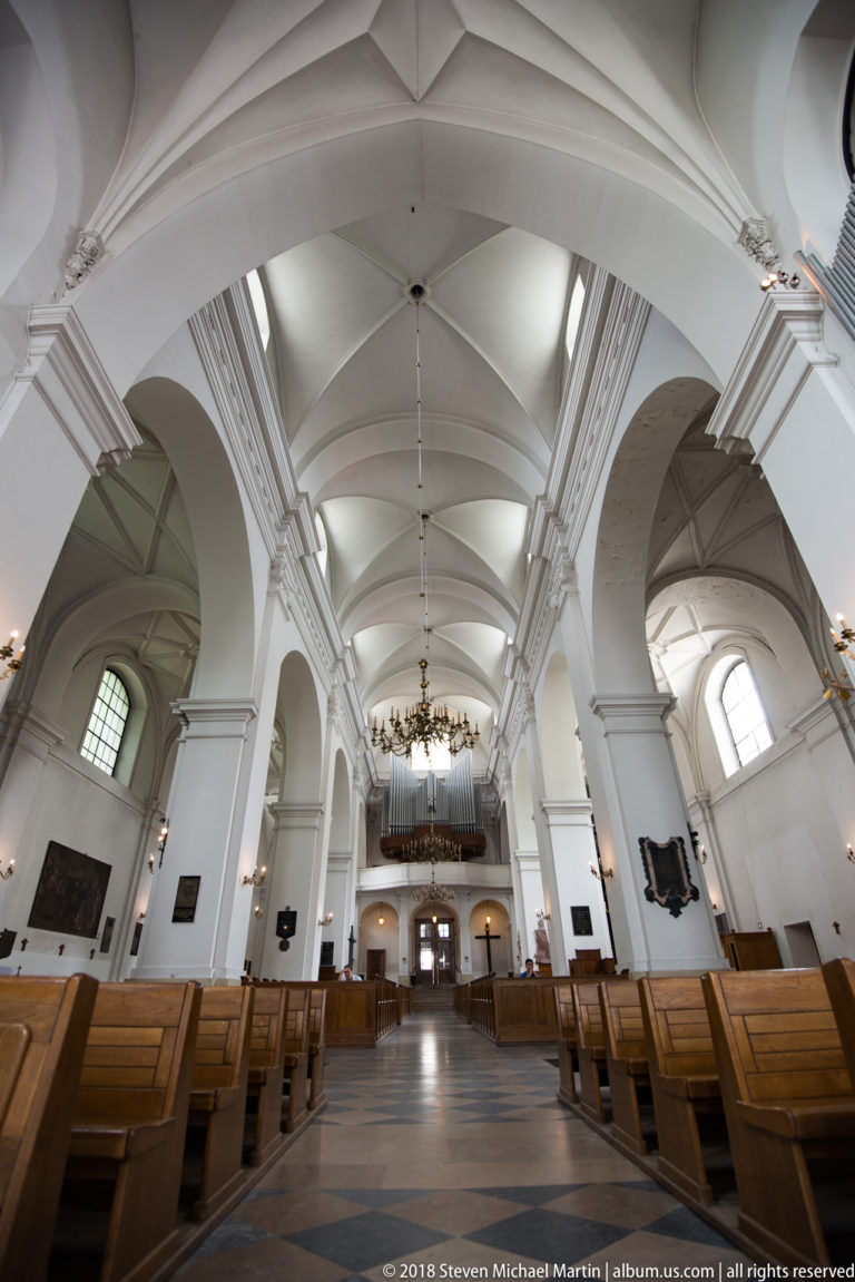 White church nave with repeated shallow vaults, wooden pews, chandeliers, side monuments, and organ loft at the rear.