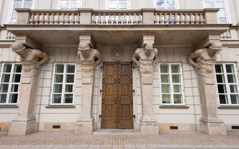 Four carved male atlantes flank a wooden doorway and support a stone balcony on a historic Warsaw palace facade.