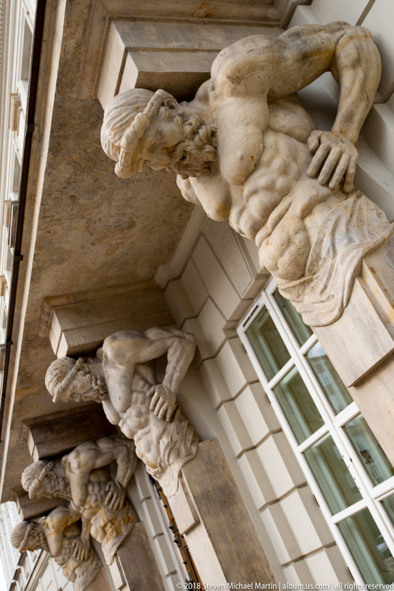 Row of muscular carved male figures serving as architectural supports beneath a projecting stone balcony.