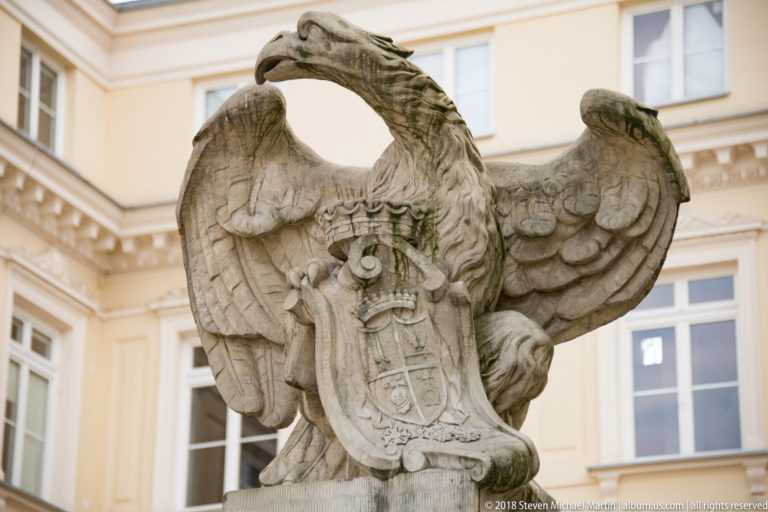 Weathered stone eagle with spread wings and crowned shield standing in a pale university courtyard.