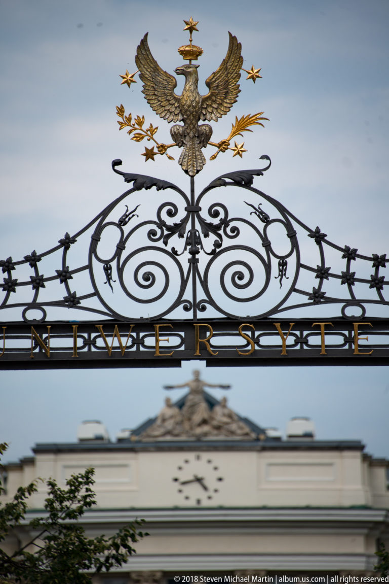 Ornamental University of Warsaw gate crest with crowned eagle, scrolling ironwork, and the word UNIWERSYTET.