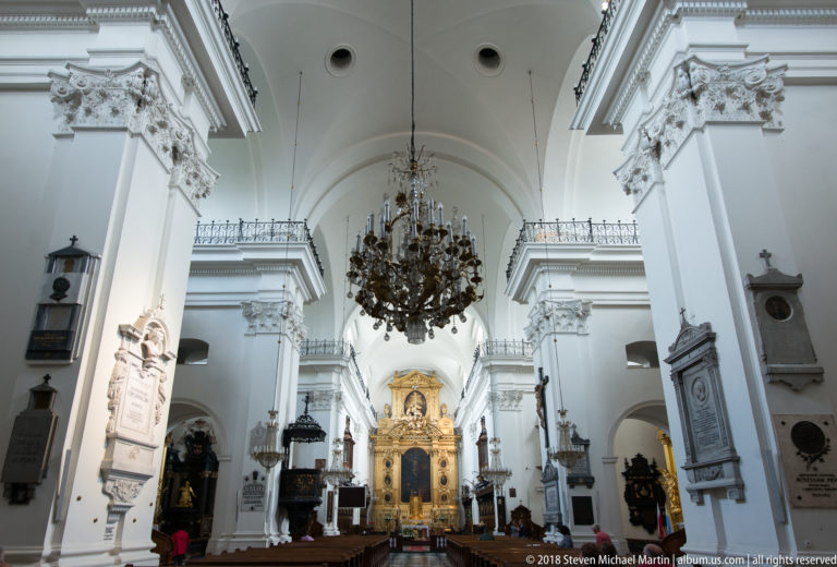 White vaulted church nave with large chandelier, wall monuments, and gilded altar centered in the distance.