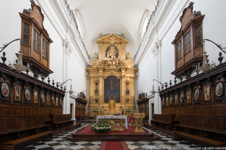 Church sanctuary with gilded high altar, dark carved choir stalls, and black-and-white patterned floor.
