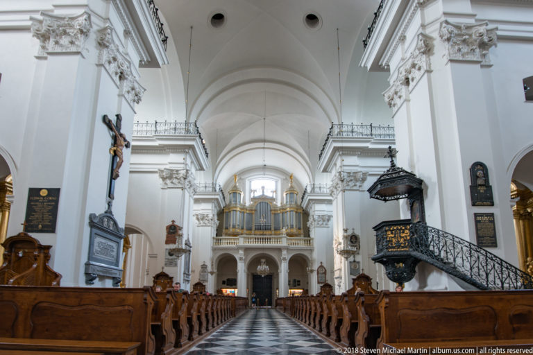 White Baroque church nave with dark wooden pews, ornate pulpit, wall monuments, and organ loft at the far end.