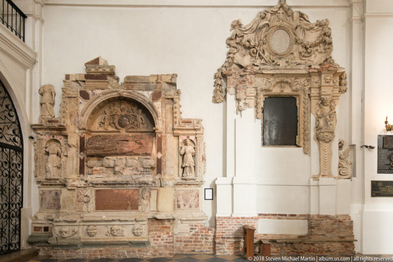 Two ornate historic wall tombs, including a reclining effigy and a richly carved vertical monument, inside a white church.