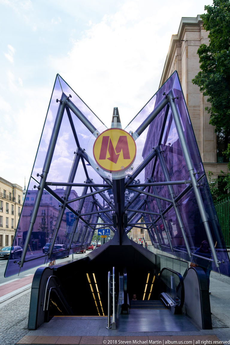 Purple angular glass canopy over a Warsaw Metro entrance with yellow-and-red M symbol.