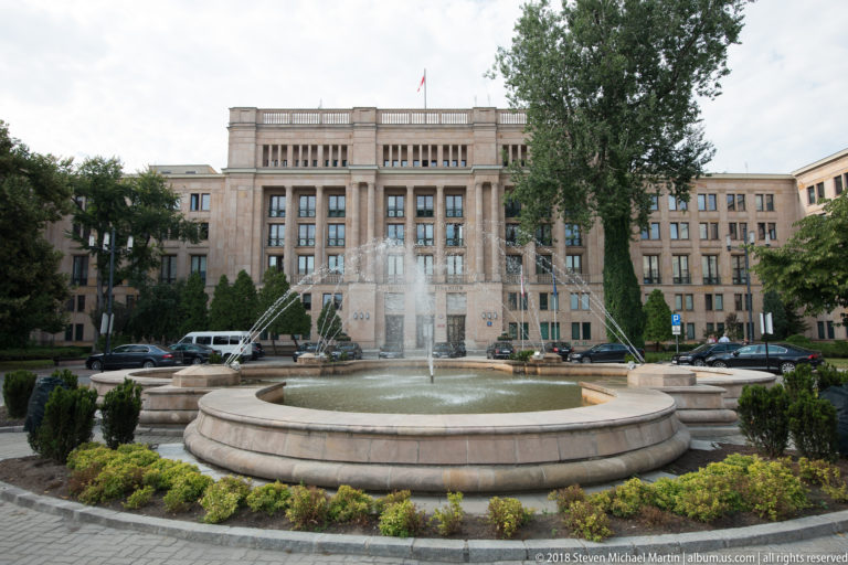 Large stone government building with tall central windows and a Polish flag behind a circular fountain and landscaping.