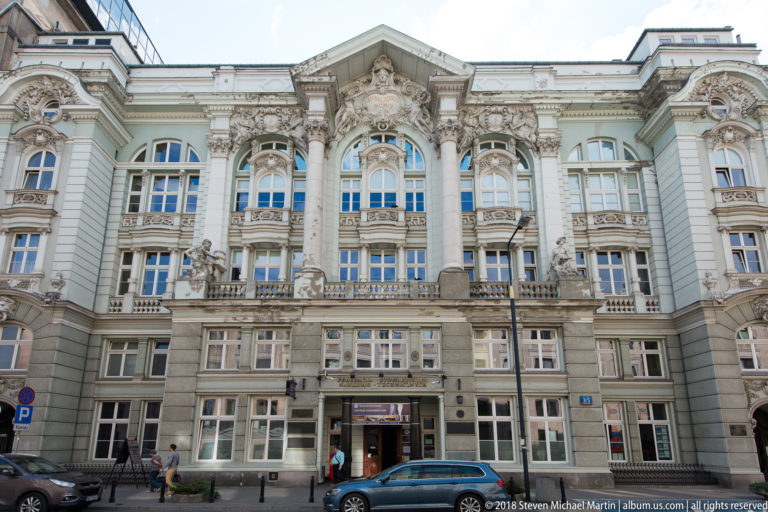 Ornate historic Warsaw facade with arched windows, paired columns, sculpted figures, balconies, and ground-floor shops.