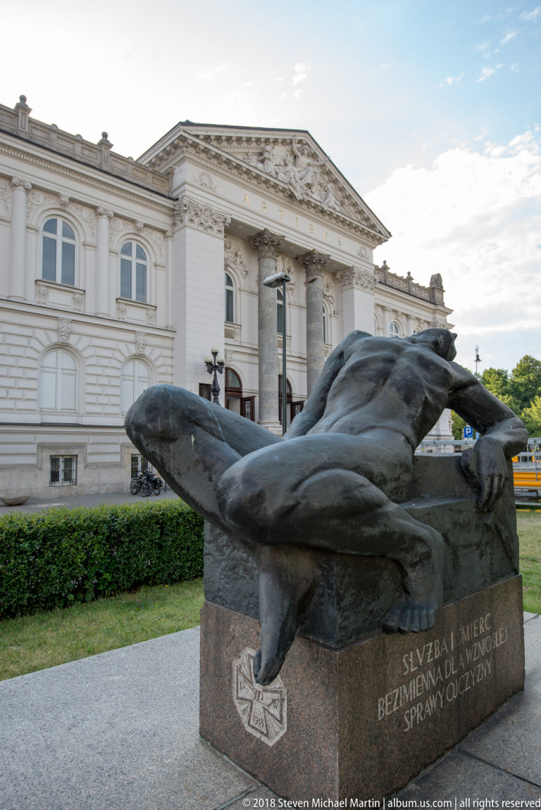 Reclining bronze human figure on a stone base with Polish inscription, set before the Zachęta gallery.