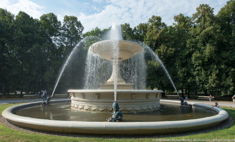 Large tiered stone fountain spraying water in a tree-lined Warsaw park with visitors around the basin.