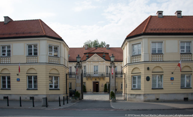 Symmetrical cream gatehouses with red roofs and Polish flags frame an entrance to a formal courtyard.