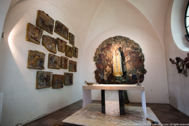Small white vaulted chapel with modern Marian shrine, bronze relief panels, and a simple stone altar table.