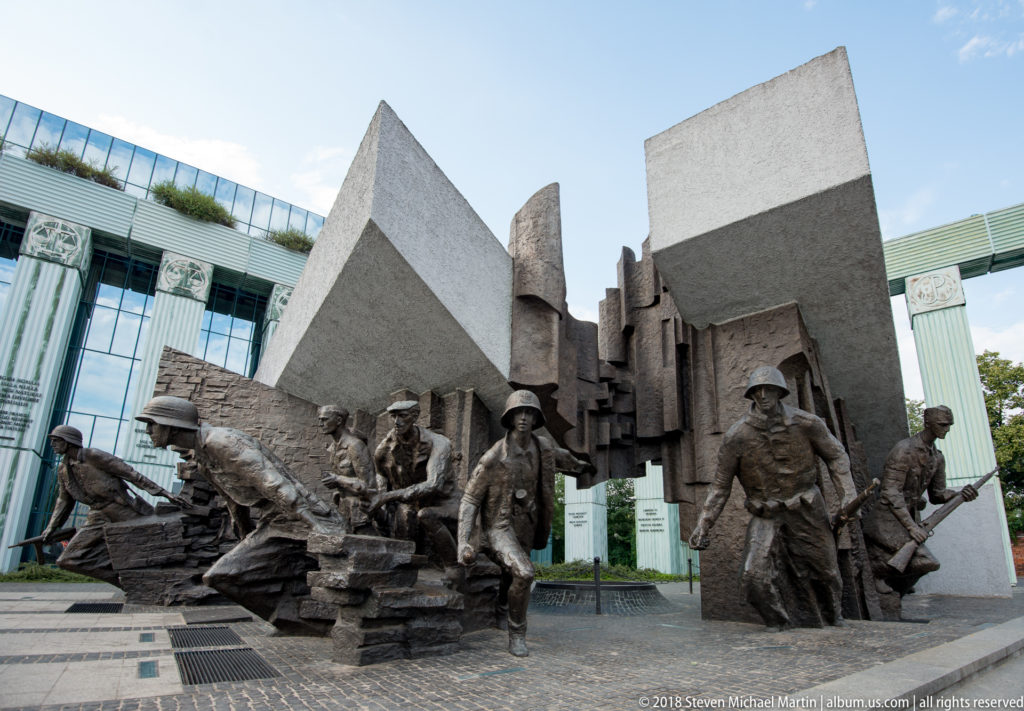 Bronze insurgent figures with helmets and weapons advance beside angular stone forms at the Warsaw Uprising Monument.