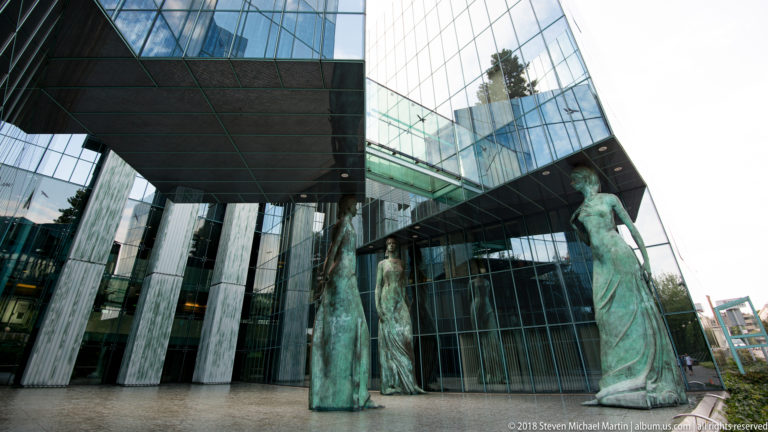 Three elongated bronze female statues stand beneath a projecting glass-and-stone modern building.