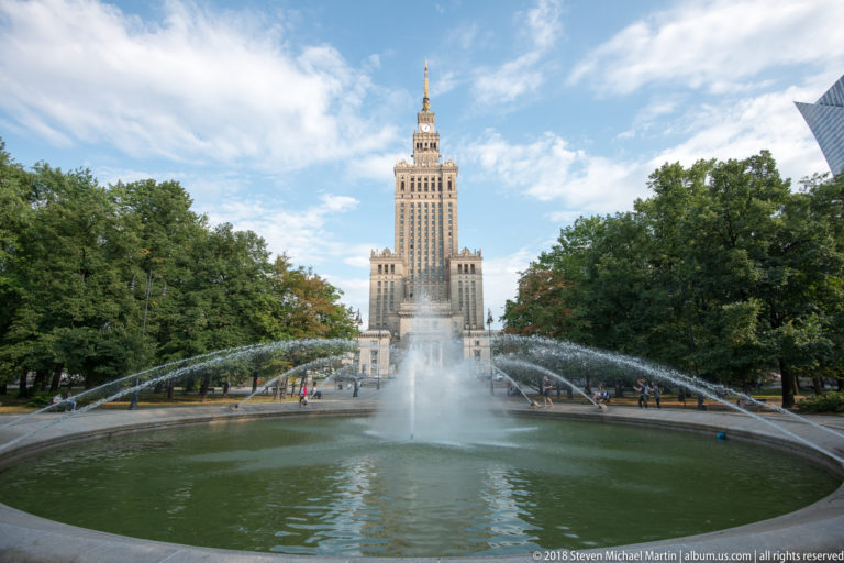 Palace of Culture and Science centered beyond a long fountain basin with arcing water jets and trees.