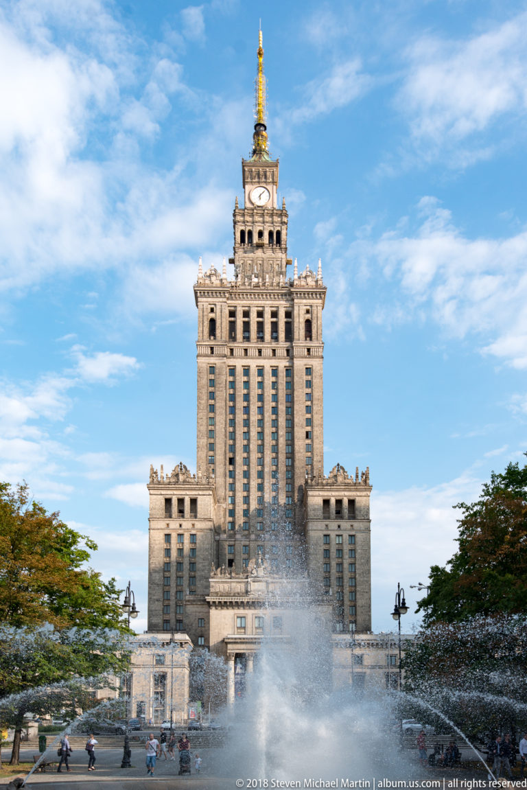 Palace of Culture and Science behind a large spraying fountain under a blue sky in central Warsaw.