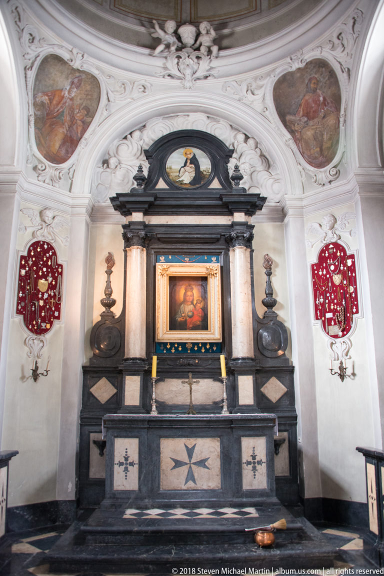 Black marble church altar with Marian painting, white eight-pointed cross, and red-and-white heraldic shields.