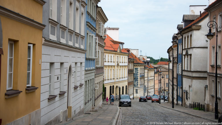 Sloping Warsaw street lined with pastel historic buildings, decorative lamps, and parked cars.