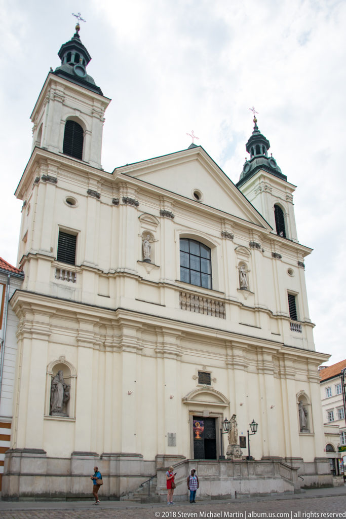 Pale Baroque church facade with twin square towers, dark cupolas, statues in niches, and a central pediment.