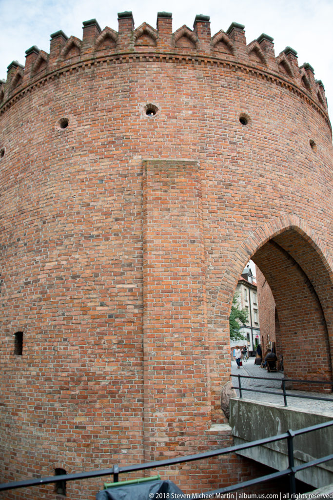 Curved red-brick Warsaw Barbican wall with crenellations, small openings, and a broad arched passage.