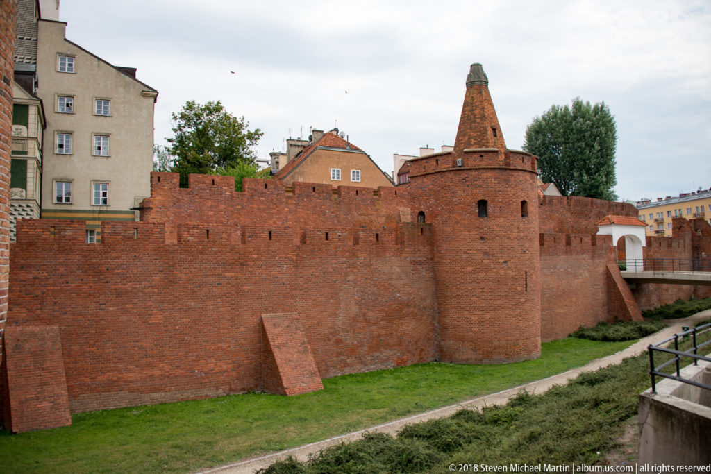 Broad view of the Warsaw Barbican's red-brick wall, projecting defensive tower, buttresses, and grassy outer ground.