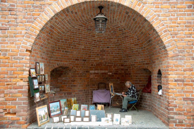 Vendor seated beneath a deep red-brick arch with framed paintings and prints displayed around the alcove.