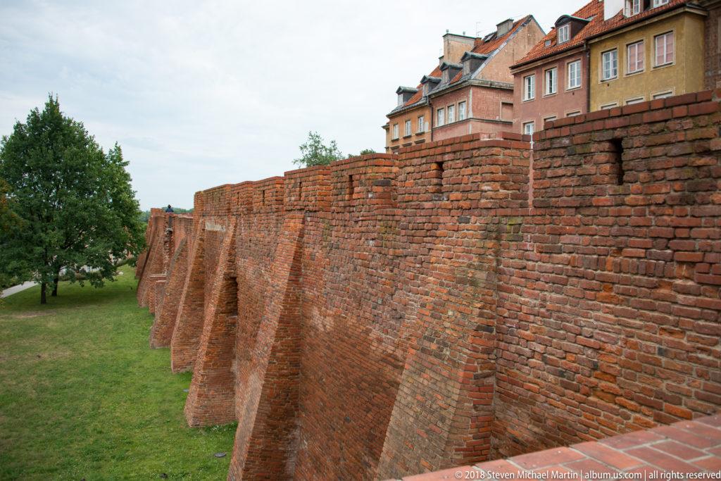 Long red-brick Old Town wall reinforced by triangular buttresses, with grass and reconstructed townhouses behind.