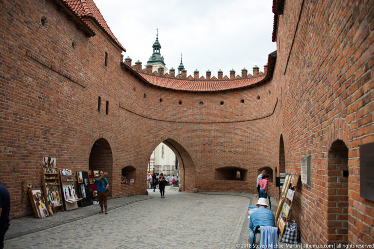 Interior of the Warsaw Barbican with high curved brick walls, pointed archway, cobbles, and art displays along the sides.