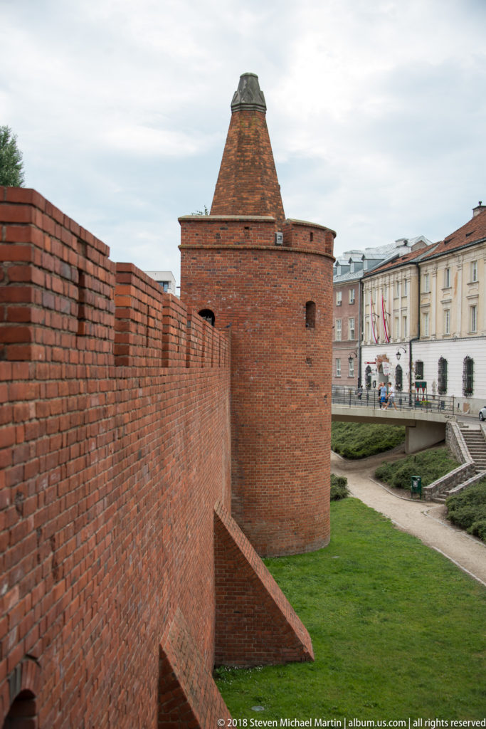 Large red-brick defensive tower joined to a curtain wall with buttresses and grassy ditch near the Warsaw Barbican.
