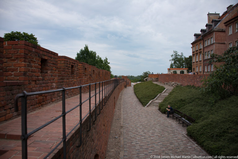 Long cobbled walkway between red-brick defensive walls, railings, planted slopes, and Old Town buildings.
