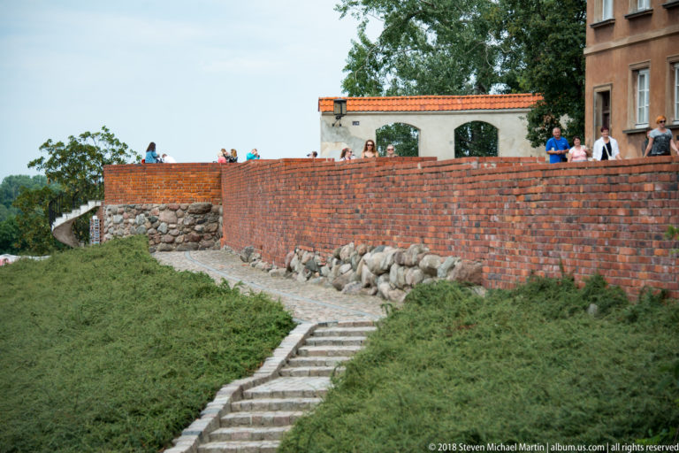 Brick defensive wall above exposed stone foundations and steps, with visitors walking along the top near Warsaw's Barbican.