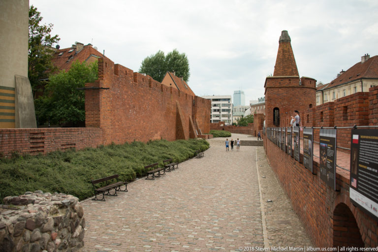 Cobbled passage between red-brick Old Town fortifications leading toward a polygonal defensive tower near the Warsaw Barbican.