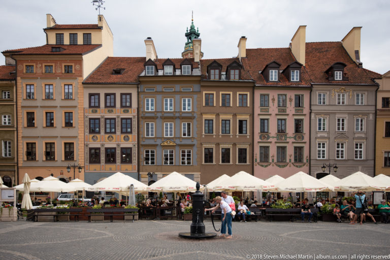 Pastel decorated townhouses and café terraces lining Warsaw's Old Town Market Square, with the mermaid fountain nearby.