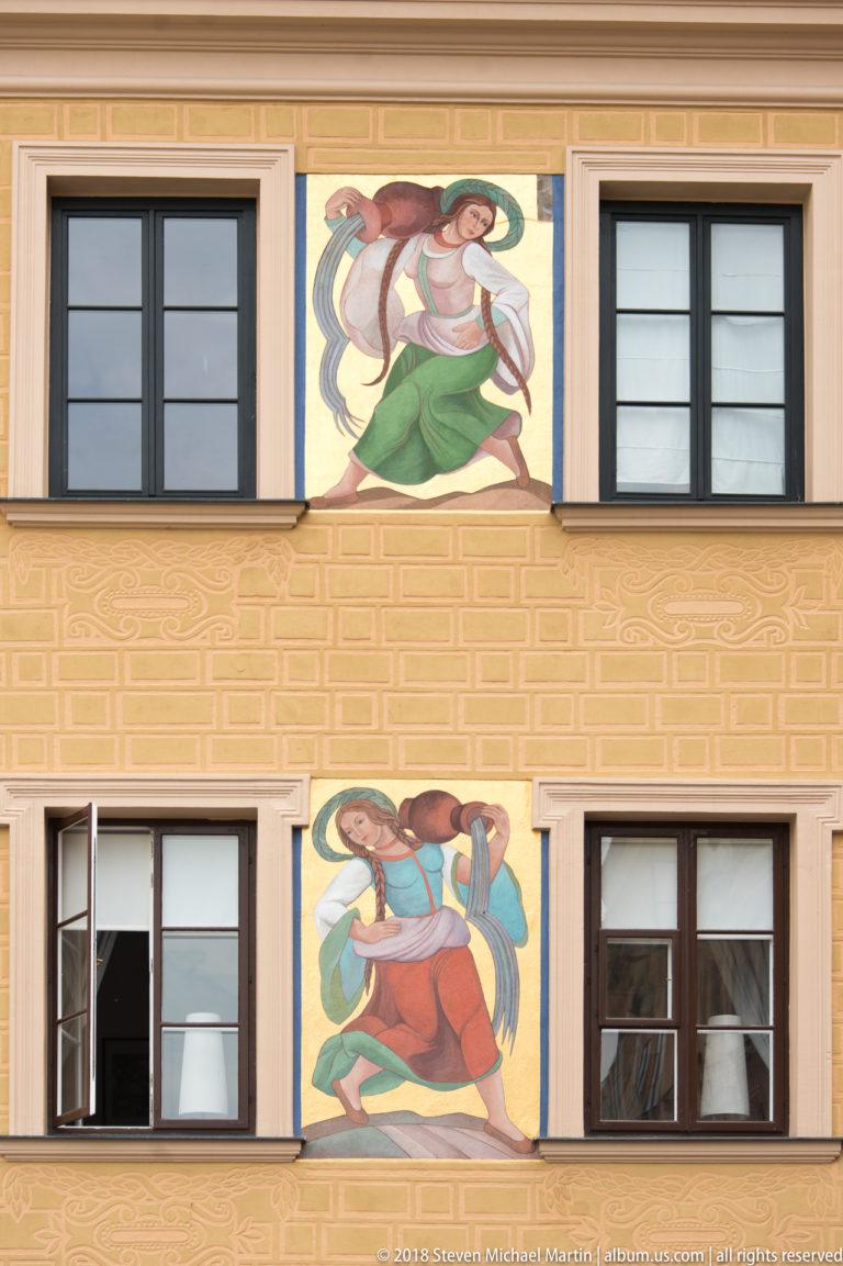 Two colorful figurative mural panels painted between black-framed windows on a pale yellow Old Town facade.