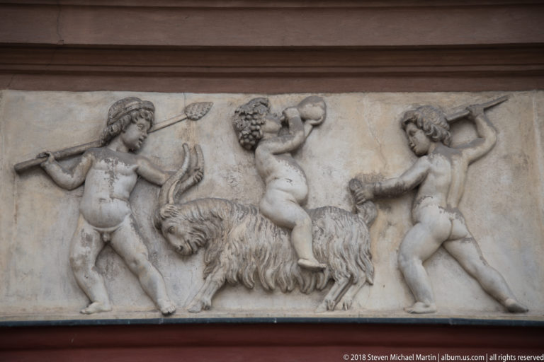 Stone relief of three youthful figures with goats, including a staff and raised vessel, on a historic facade.