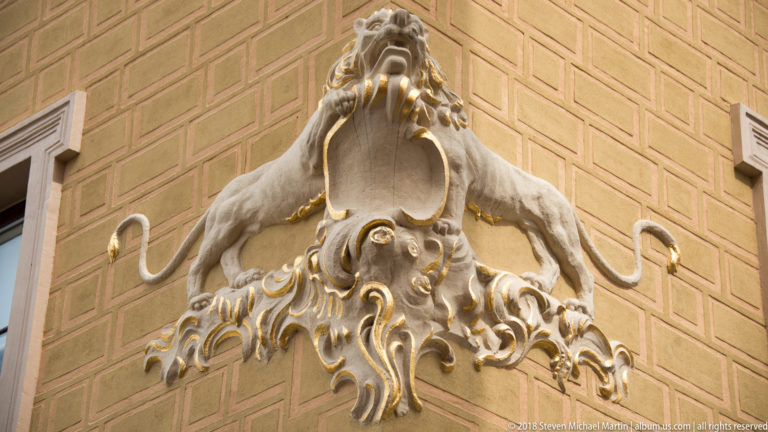 Grotesque lion-like creature above a shield and curling sea-creature forms with gilded accents on a yellow facade.