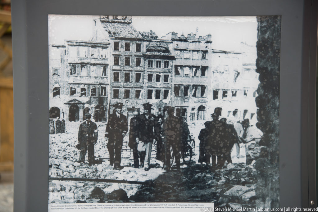 Historic black-and-white photograph of people standing amid rubble and damaged buildings in wartime Warsaw.